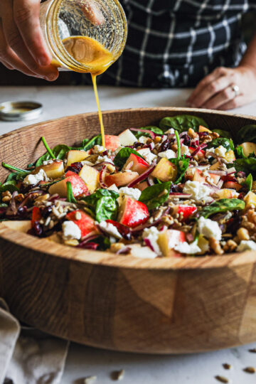 Spinach salad with apples, walnuts, dried cranberries, sunflower seeds, goat cheese, and apple cider vinaigrette being drizzled over the top.