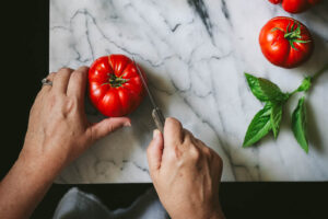 Overhead view of woman's hands preparing a ripe tomato on a marble surface with fresh tomatoes and basil.