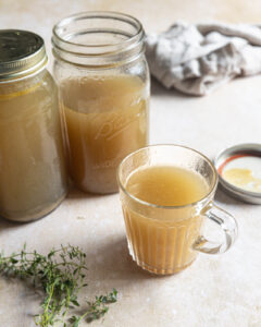Homemade chicken bone broth served warm in a glass mug with jars of broth behind it.
