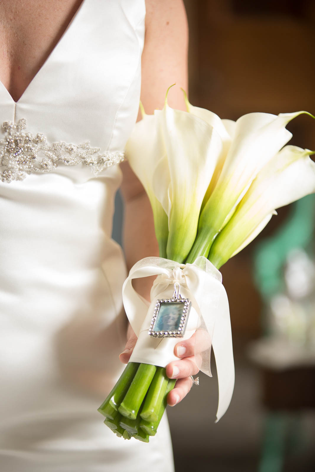 Bride holding a bouquet of white calla lilies with a small, framed photograph of her mother on her wedding day tied to the stems.