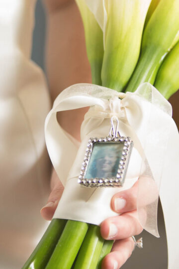 Bride holding a bouquet of white calla lilies with a small framed photograph of her mother on her wedding day tied to the stems.